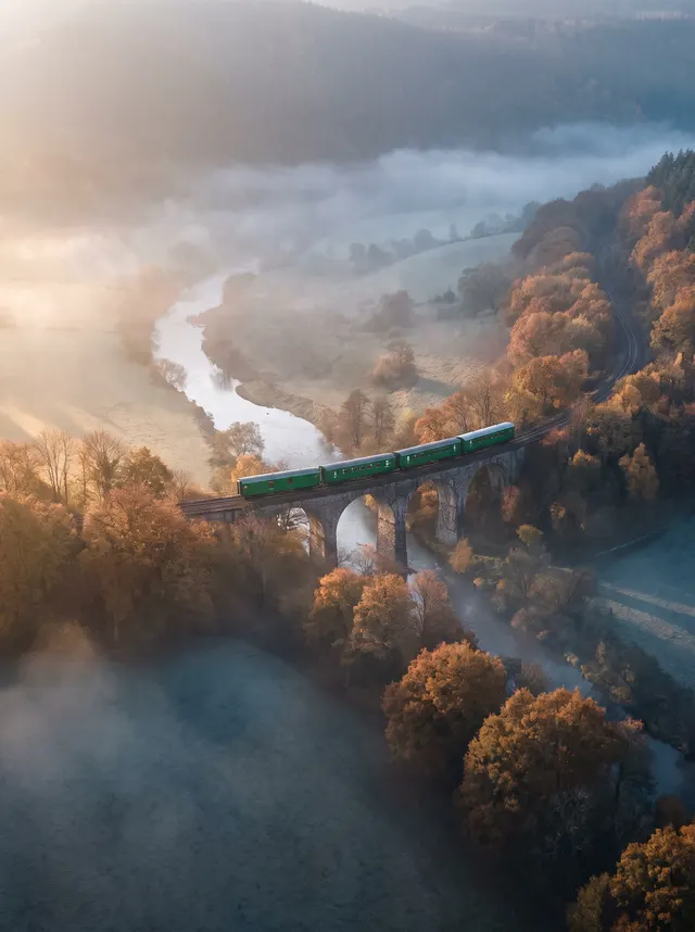 Green train crossing a stone viaduct over an autumn river valley in mist